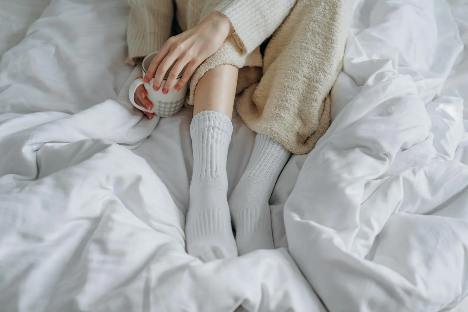 An adult enjoying a warm drink while relaxing in a cozy bed with a knitted blanket and white socks.