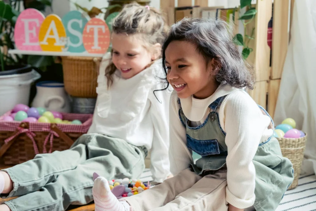 Positive multiracial children in casual clothes sitting on floor in light room with colorful eggs and decorations during Easter holiday