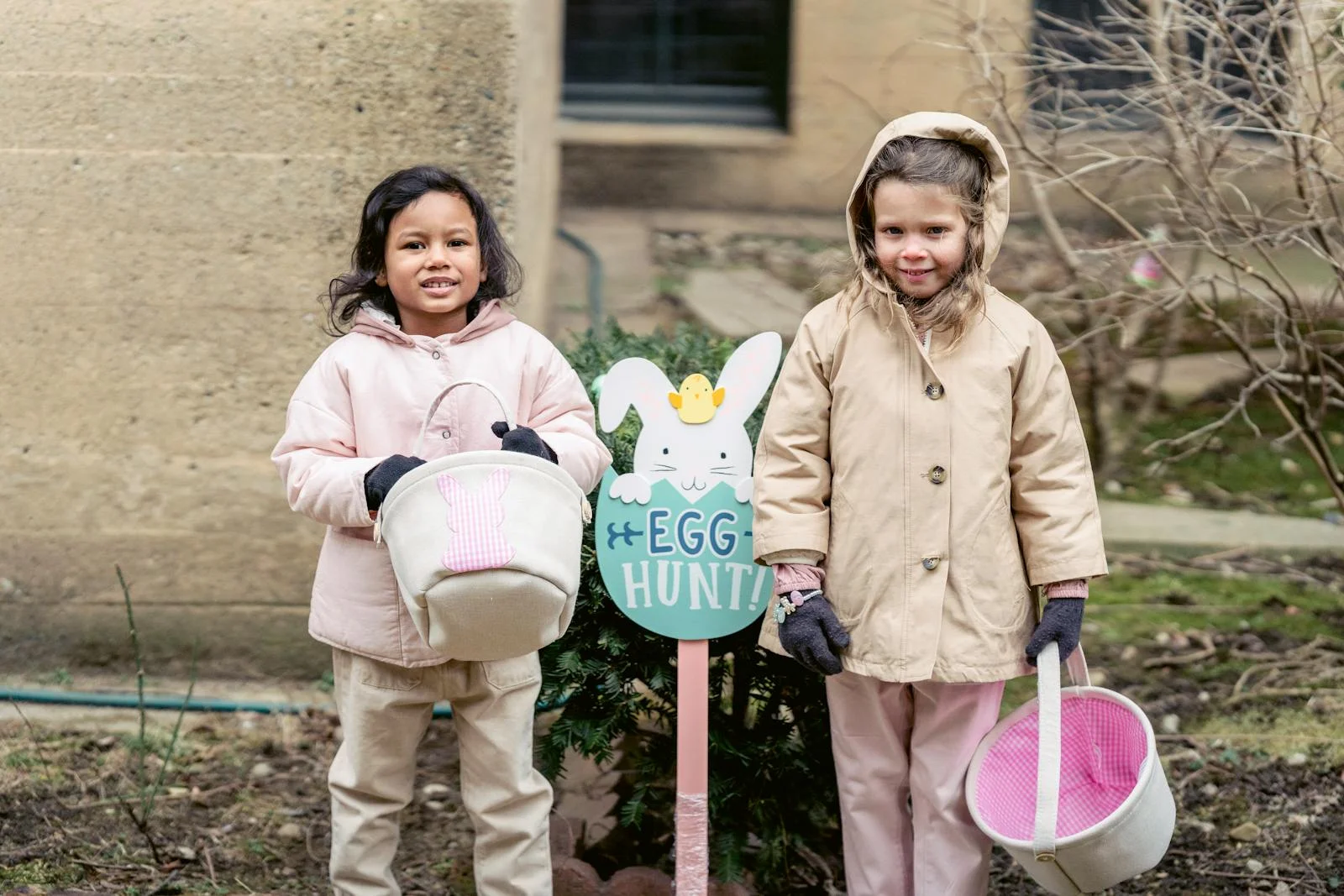 Two children in warm clothes smiling during an outdoor Easter egg hunt, holding baskets and standing by a sign.