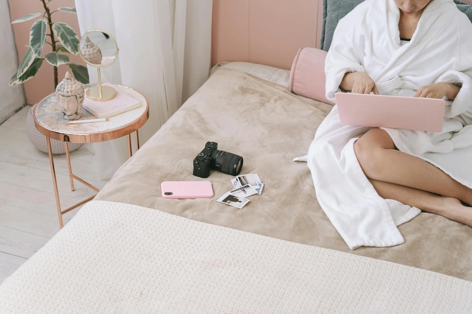 Woman in a robe using a laptop on a bed with a camera and photos nearby.