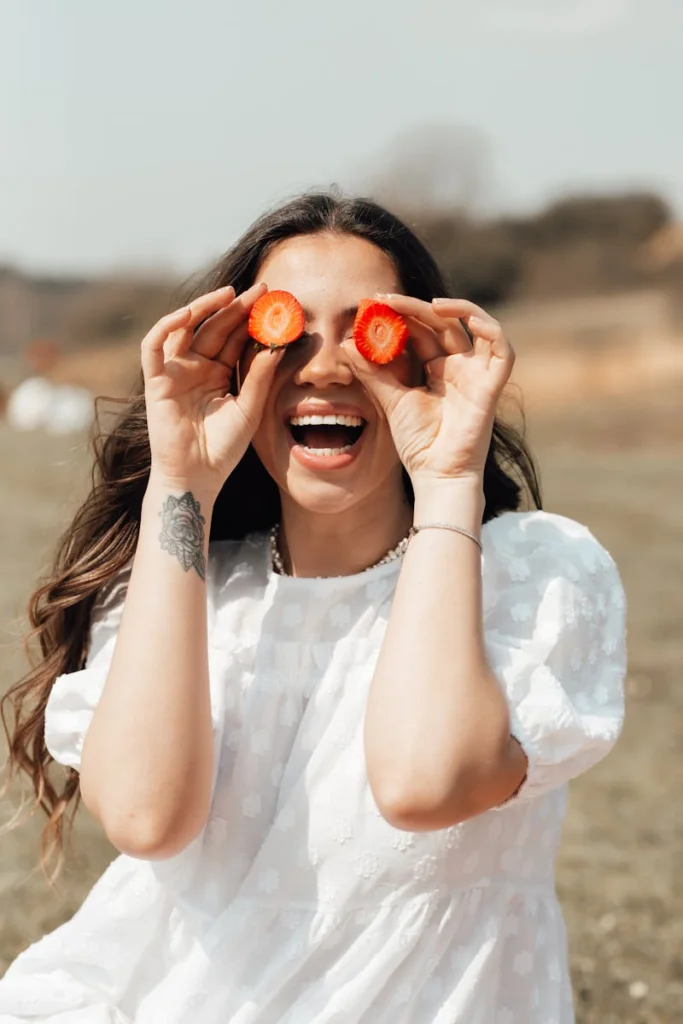Smiling woman holding strawberry slices over eyes in a sunny outdoor setting.