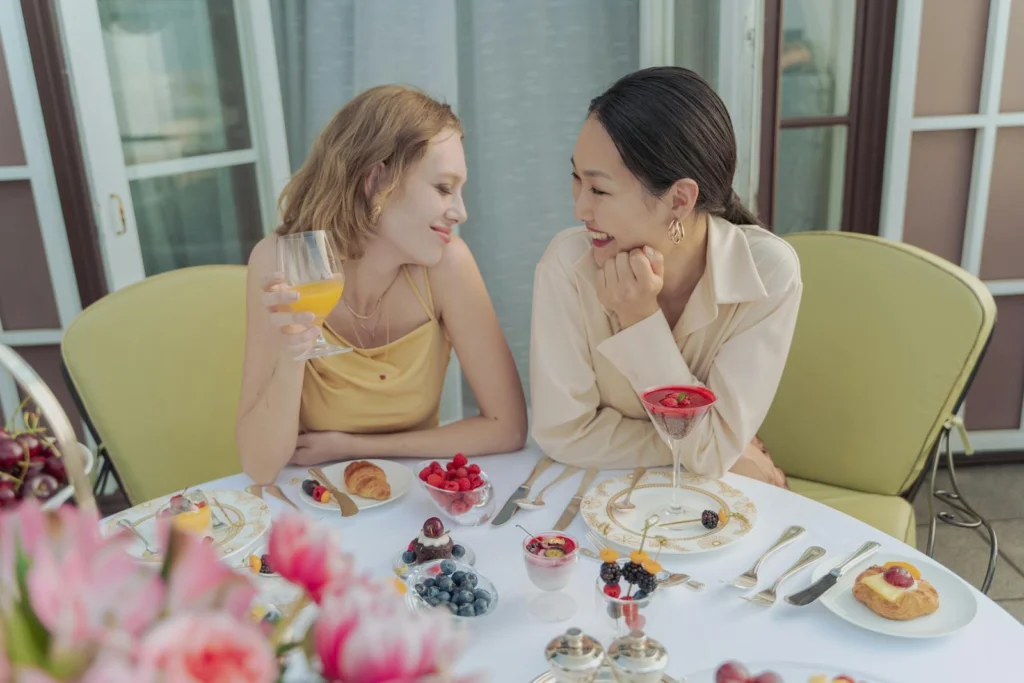 Two women sharing a joyful breakfast with berries and drinks, outdoors on a sunny day.