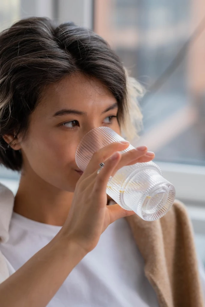 Asian woman sipping water indoors, capturing a moment of refreshment and relaxation.