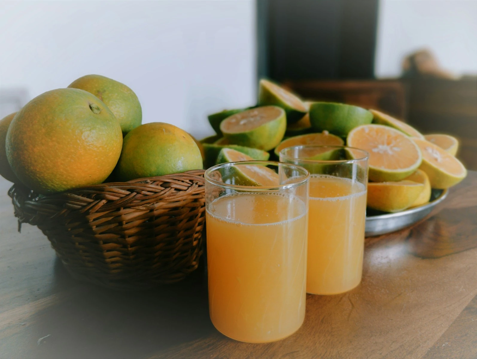 two glasses of orange juice next to a basket of oranges