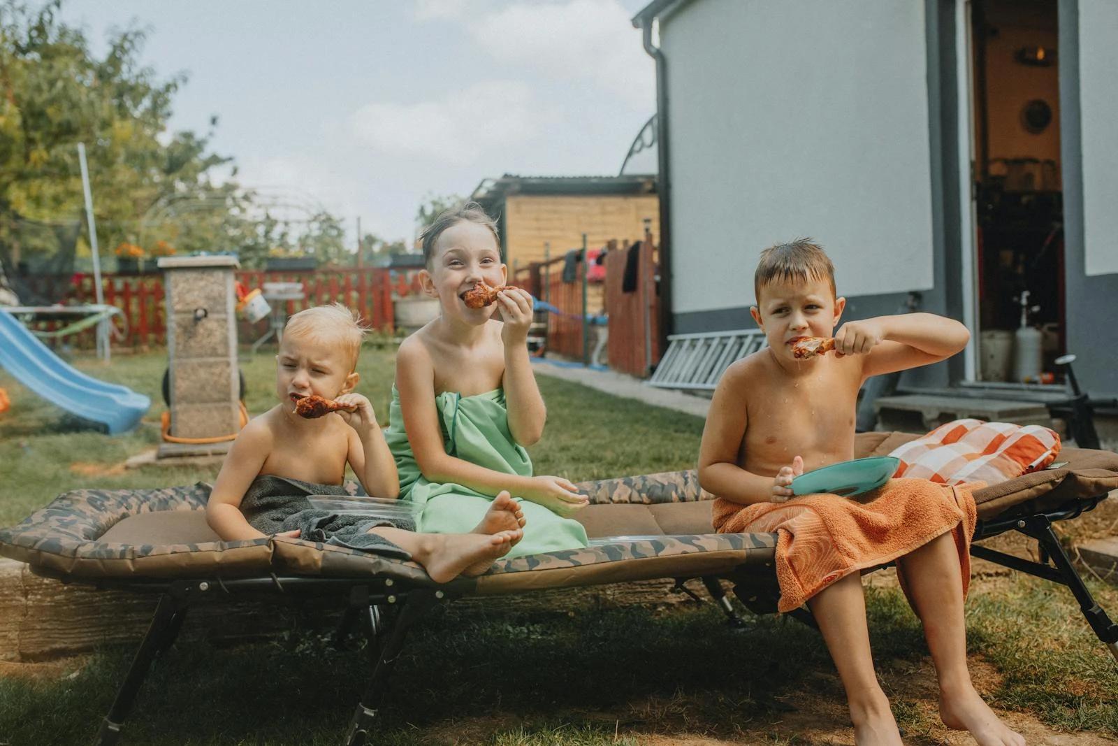 Three children laughing and eating snacks outside during a warm summer day.