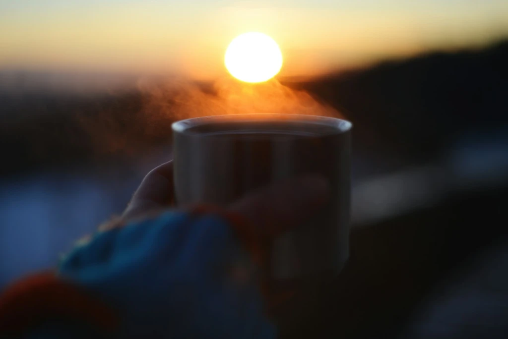 A person holding a coffee cup with the sun setting in the background