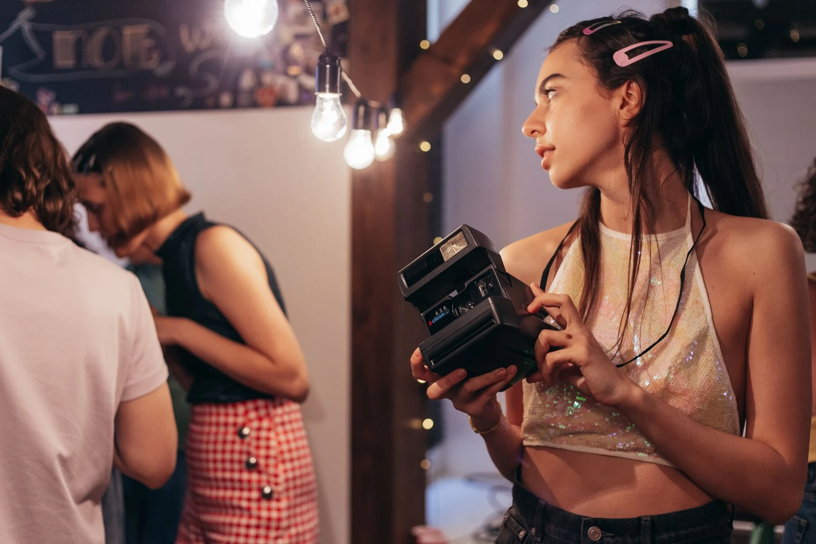 A young woman with long hair holds a polaroid camera at a casual indoor gathering with friends.