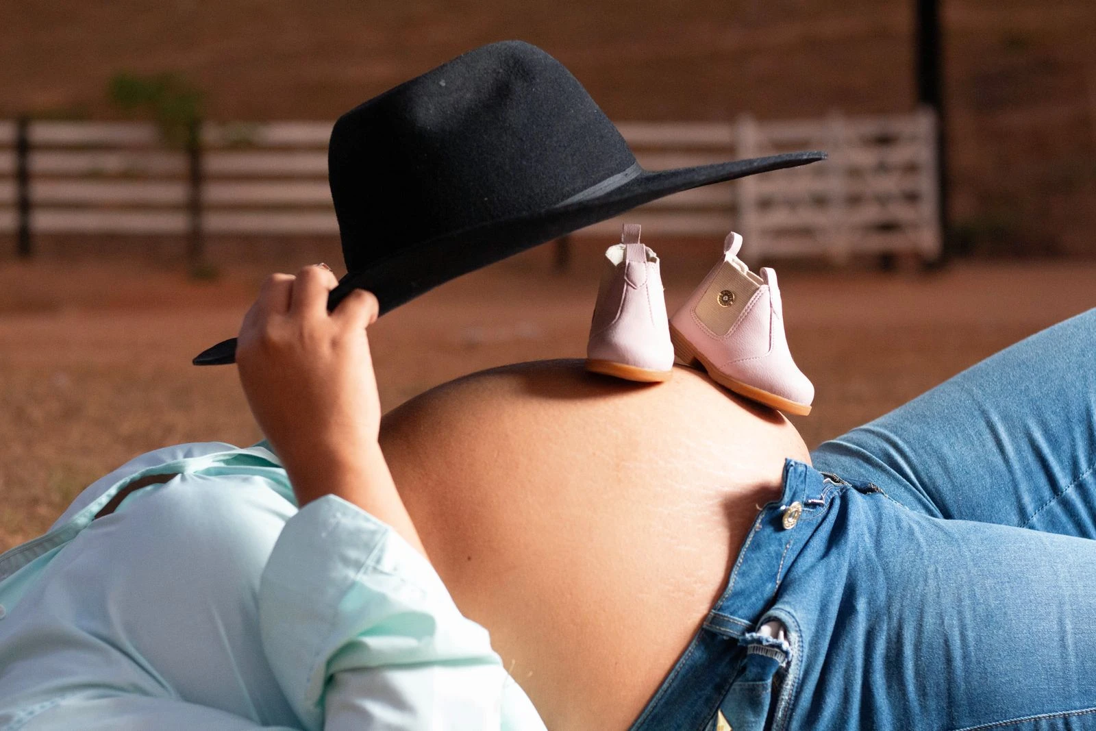 Pregnant belly with baby shoes on top and woman in hat, outdoors.