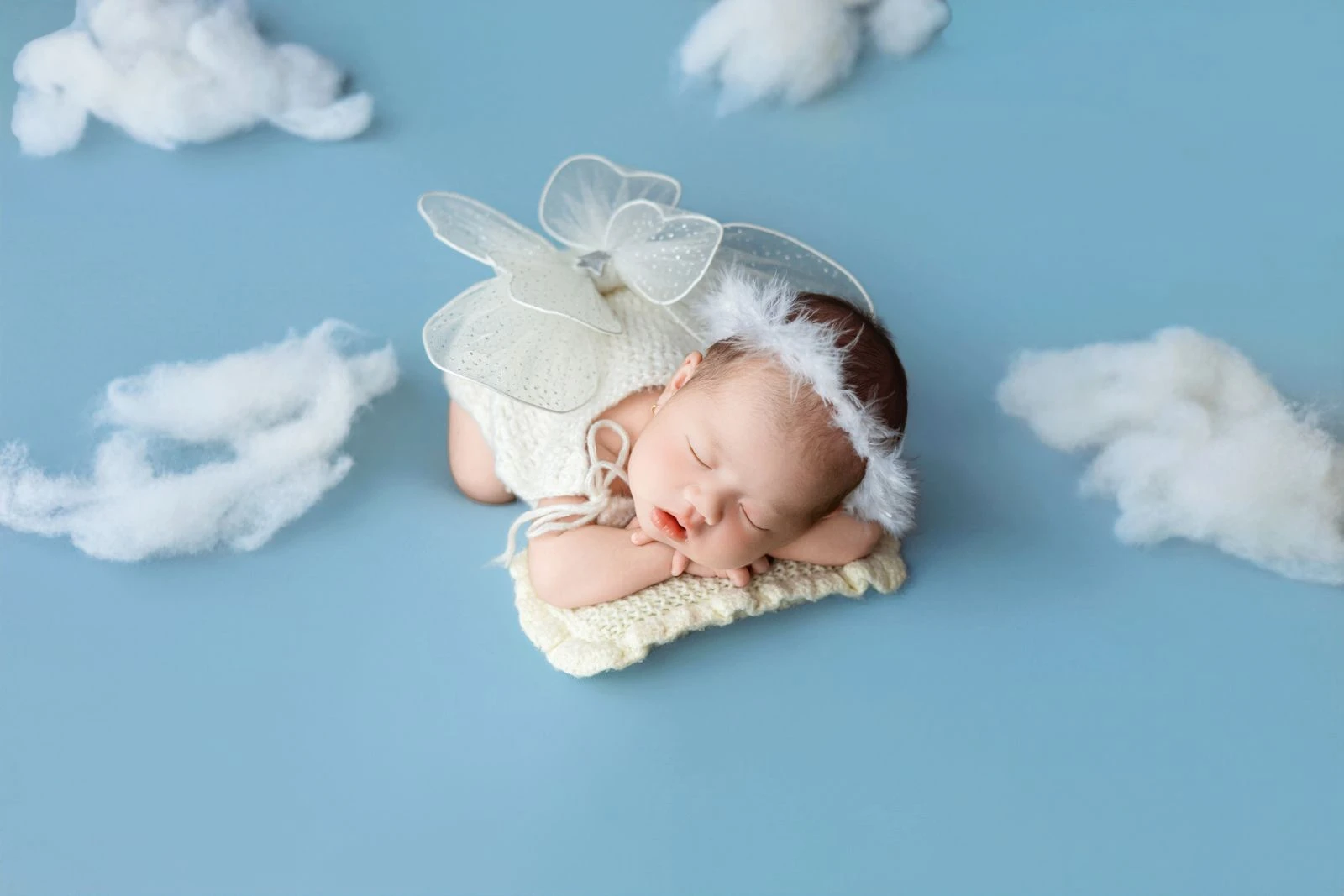 Cute newborn dressed as an angel sleeps on a cloud-like background in a studio setting.