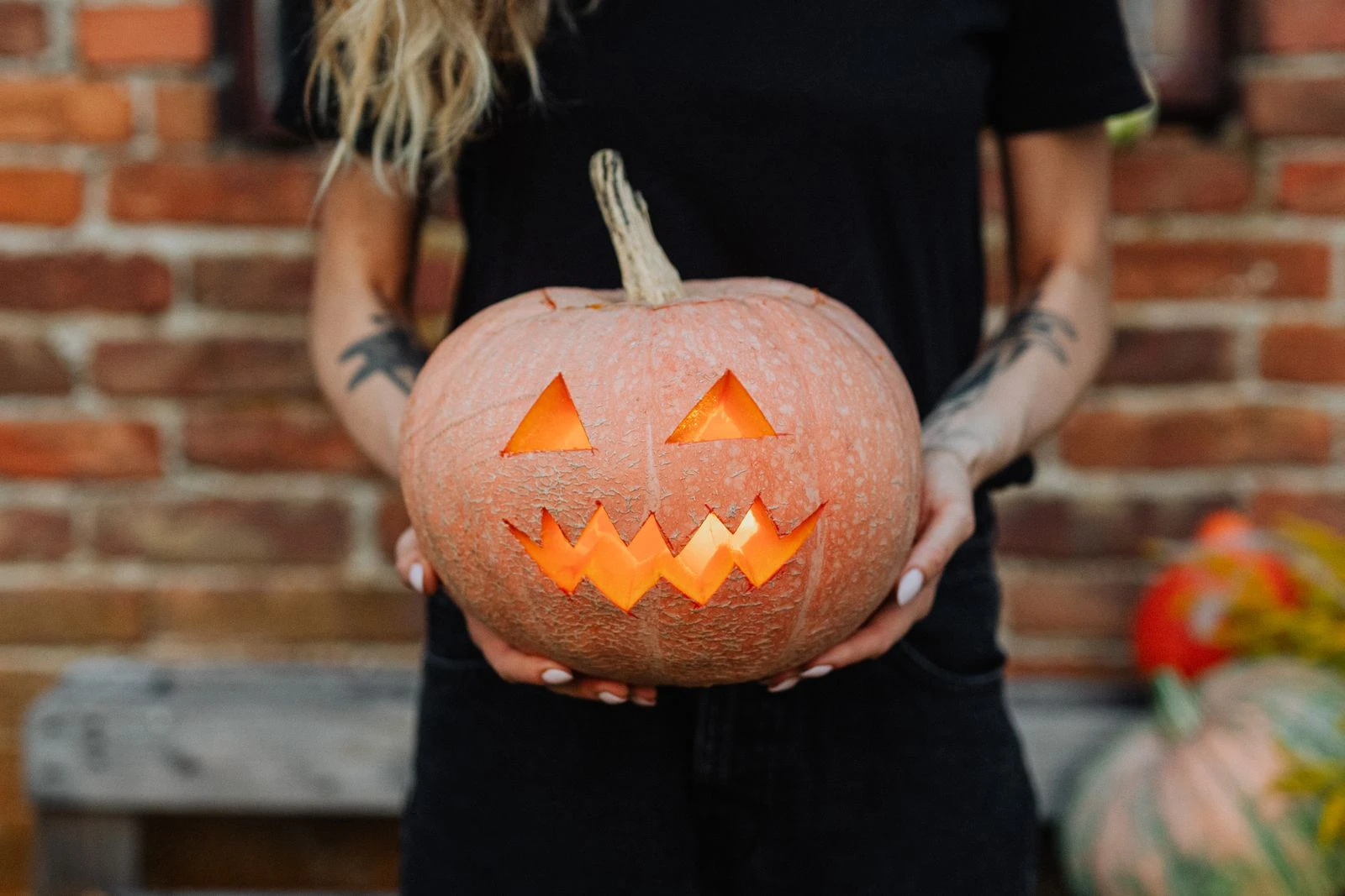 A woman with tattoos holds a glowing jack-o'-lantern in a rustic outdoor setting, perfect for Halloween-themed imagery.