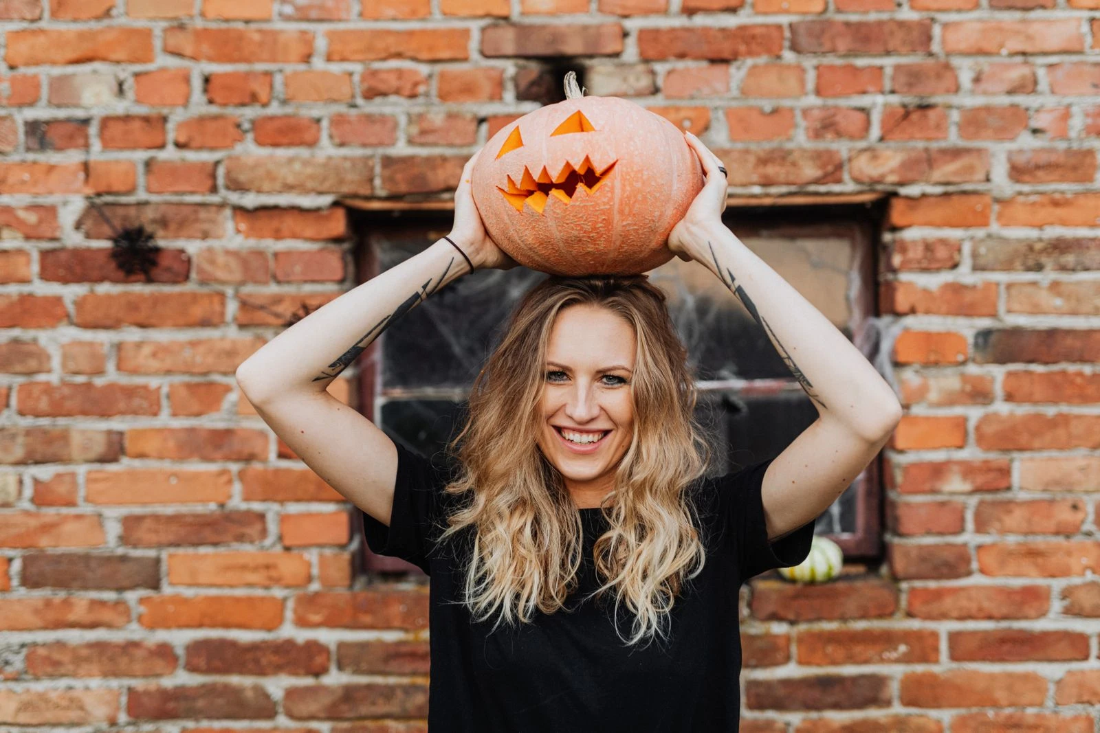 Woman holding a carved pumpkin over her head, smiling in front of a brick wall.