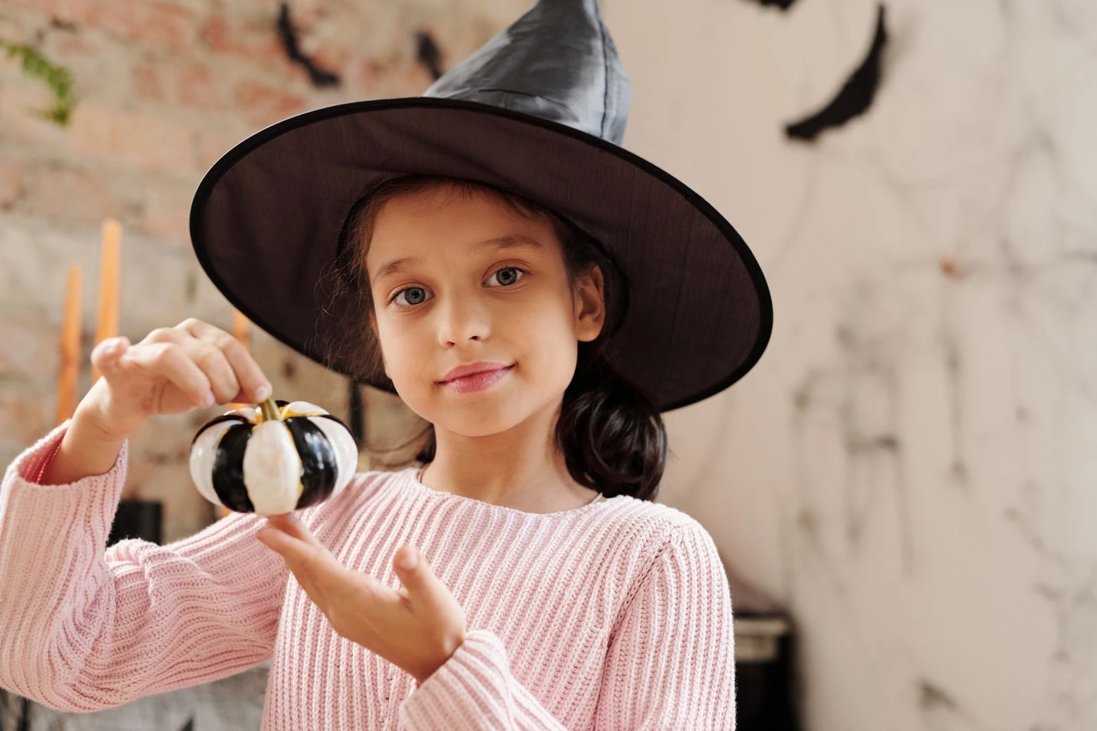 A young girl in a witch costume holding a decorative pumpkin indoors for Halloween.