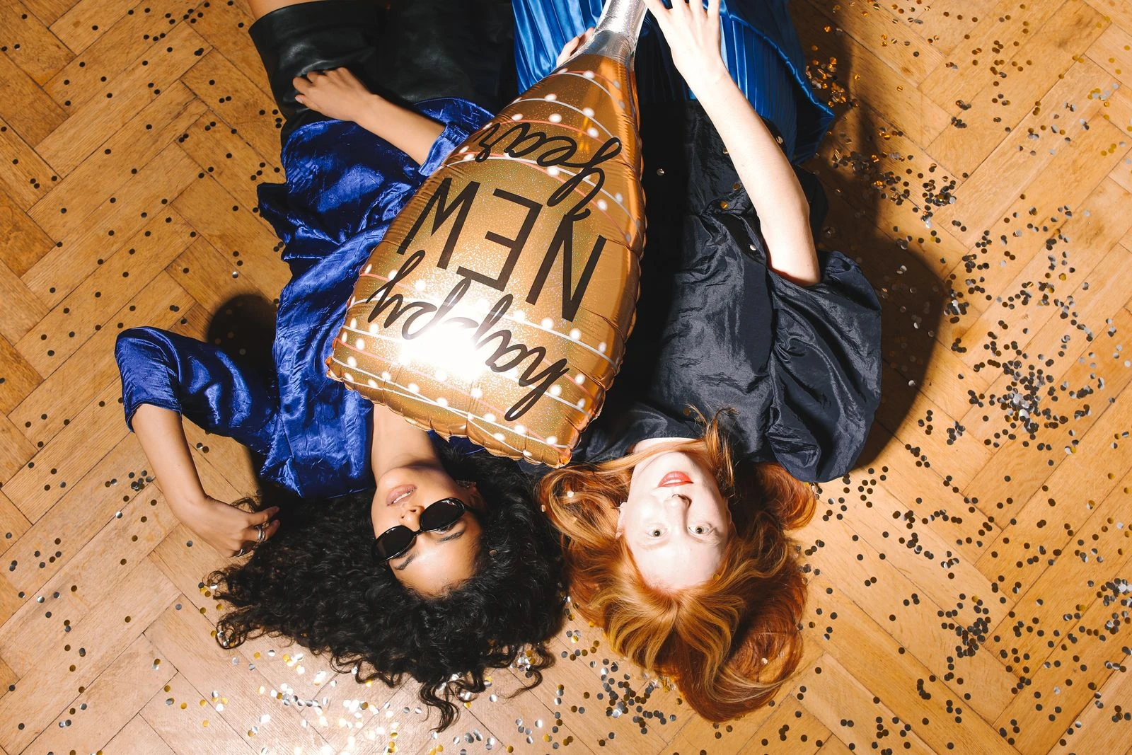 Two women celebrating New Year with a balloon and confetti on a wooden floor.