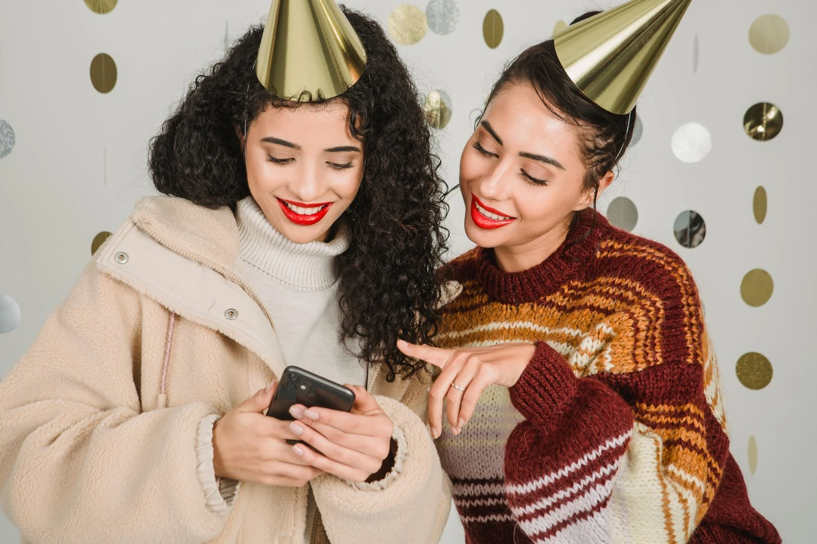 Content female friends wearing party hats surfing cellphone and standing on white background with hanging garland during party in studio