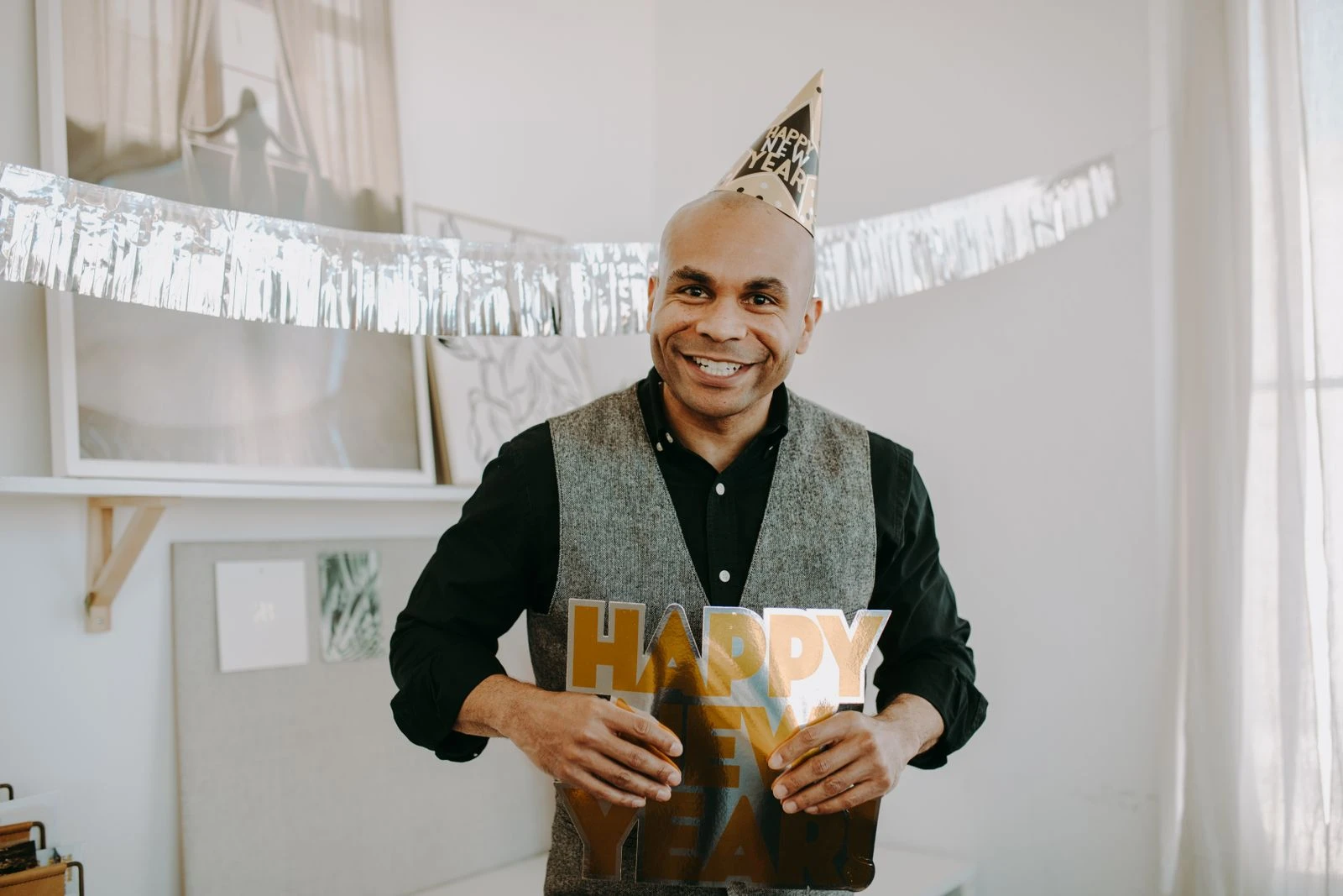 Smiling man holding Happy New Year sign during indoor celebration, with festive decorations.