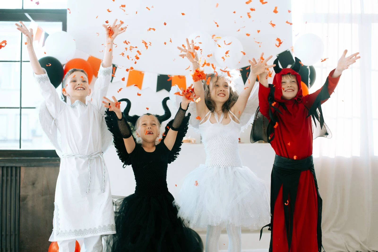 Children in angel and demon costumes celebrating Halloween indoors with confetti and balloons.