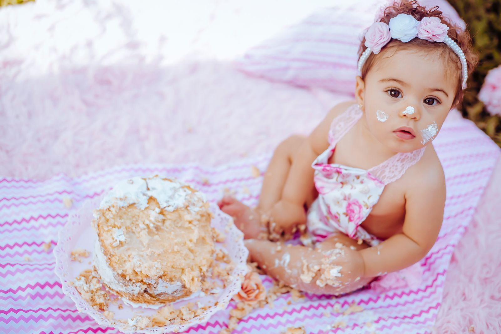 Cute baby enjoying a cake smash for her first birthday, sitting outdoors on a blanket.