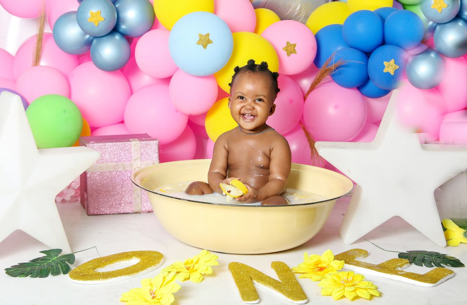 Smiling baby in a tub with colorful balloons and decorations for a first birthday celebration.