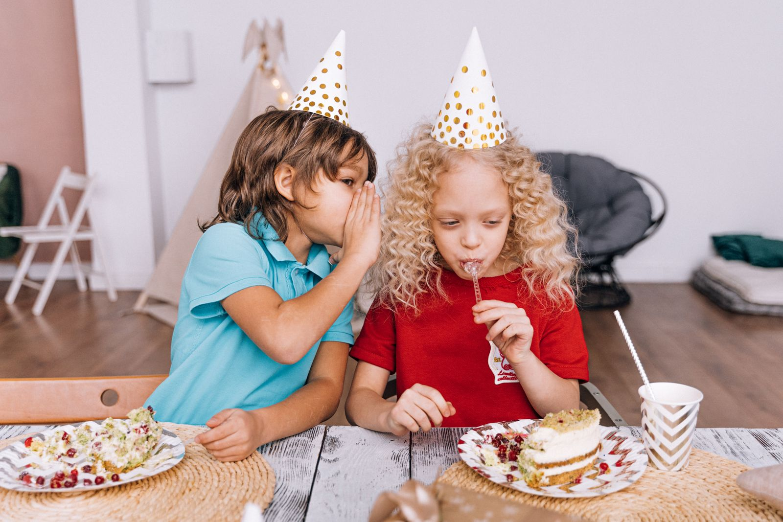 Two children at a birthday party whispering and enjoying cake indoors.