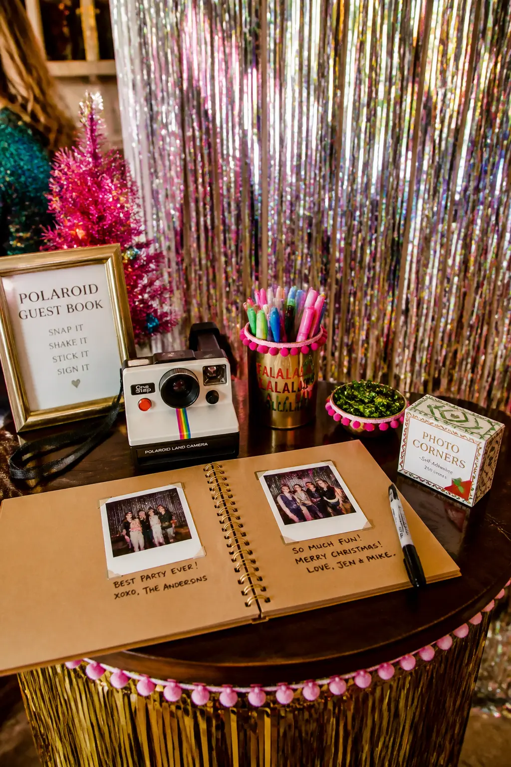Polaroid Print Station — An instant print camera on a small table beside the tinsel backdrop, with a stack of adhesive photo corners and a kraft paper