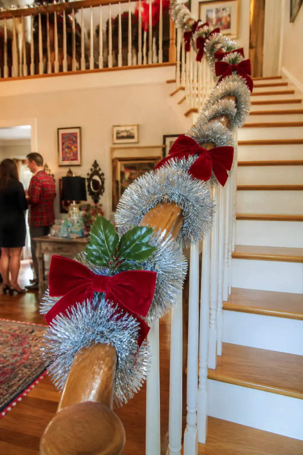 Tinsel Swag Banister — A staircase banister wrapped in silver tinsel garland with sprigs of artificial holly tucked in at intervals and red velvet bows tied