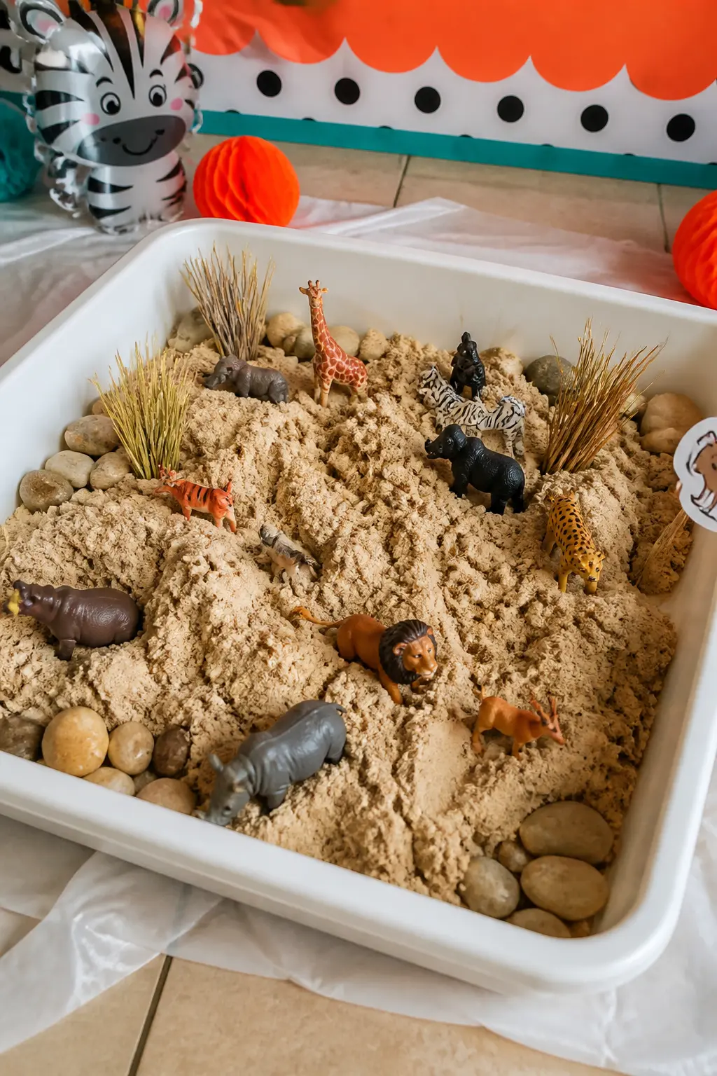 Animal Habitat Sensory Bin — A wide shallow bin filled with kinetic sand in savanna tan, small plastic safari animals half-buried in it and a scatter of dried