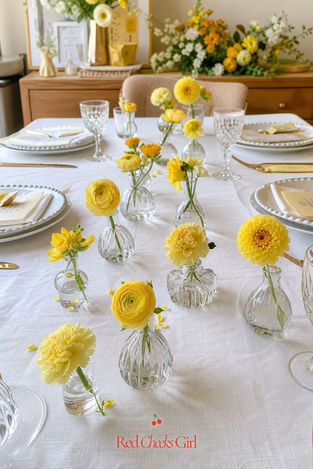 Butter Yellow Bud Vase Trail — A dining table set with a scattered trail of small glass bud vases, each holding one or two butter yellow blooms against white