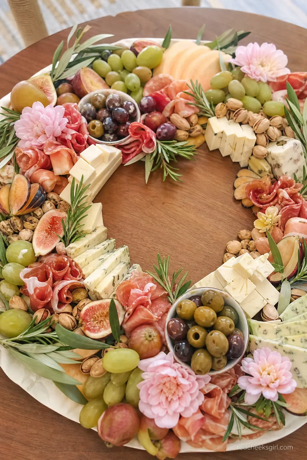 Olive and Blush Grazing Board Wreath — A large round grazing board arranged in a wreath shape, with olive colored herbs and blush colored fruit filling in the