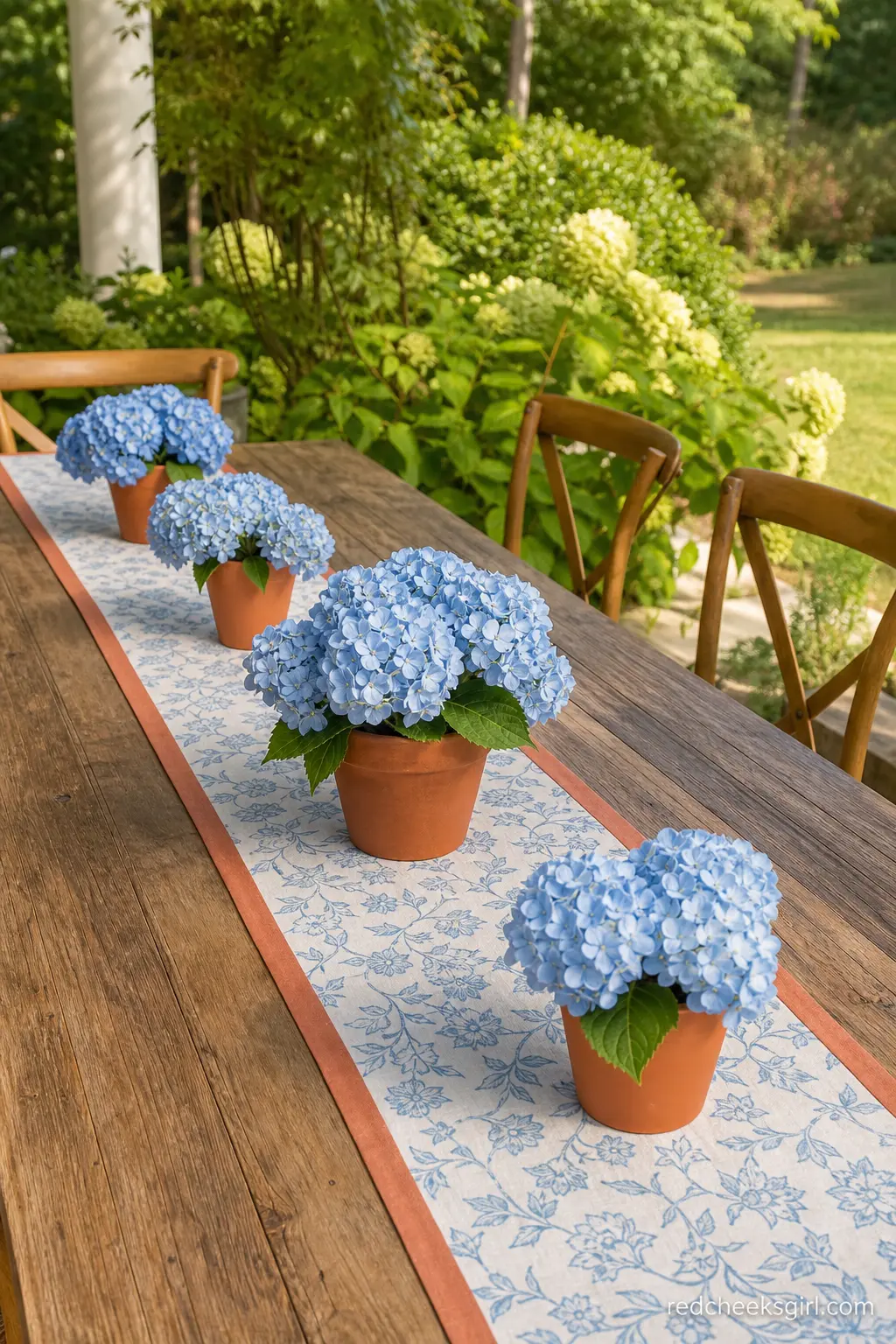 Terracotta and Powder Blue Table Runner — Down the length of an outdoor table, a row of small terracotta pots holding powder blue hydrangea heads sits directly