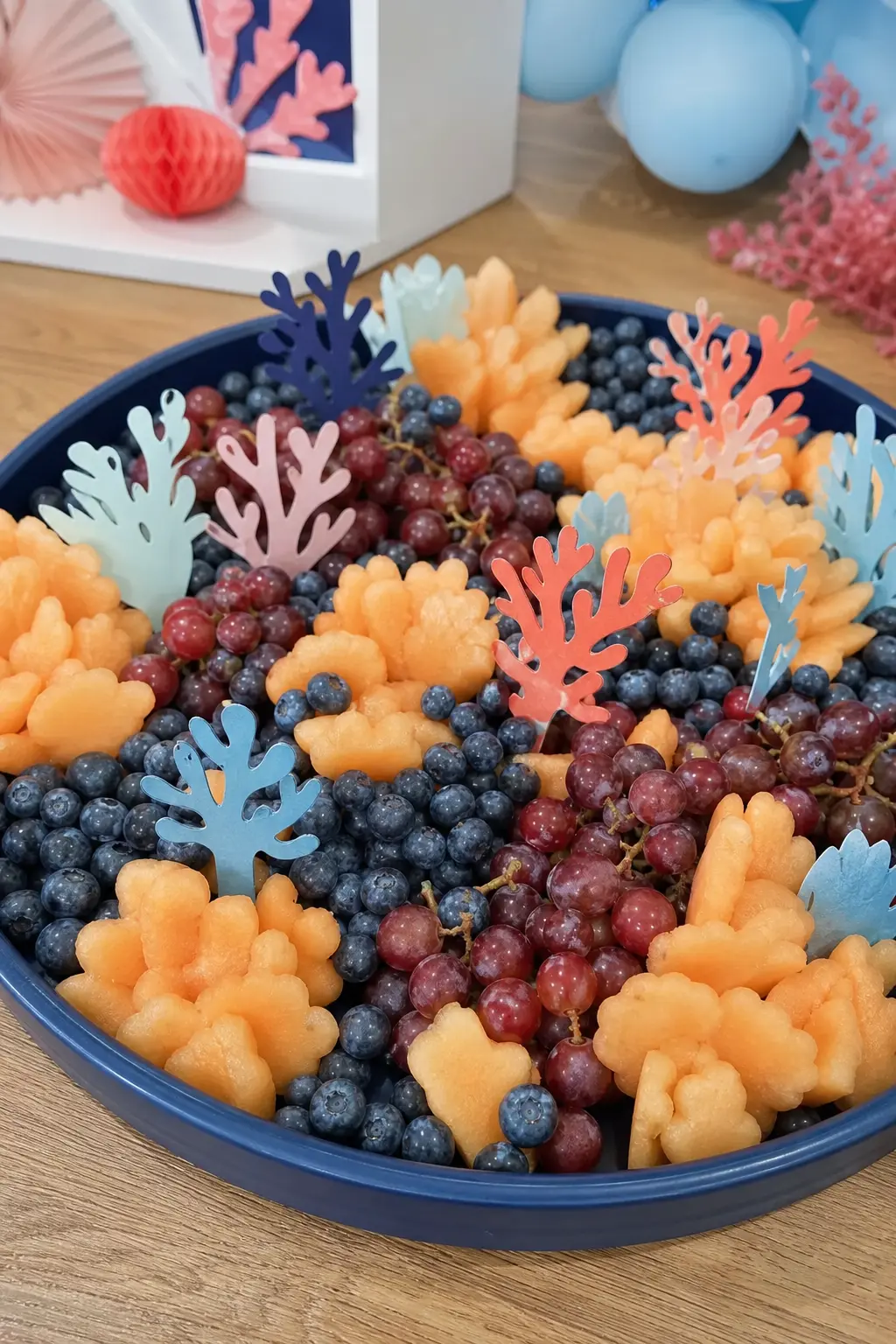 Coral Reef Grazing Board — A large navy serving board, fruit arranged in blue and coral tones with a few standing paper coral picks, turns a fruit platter into