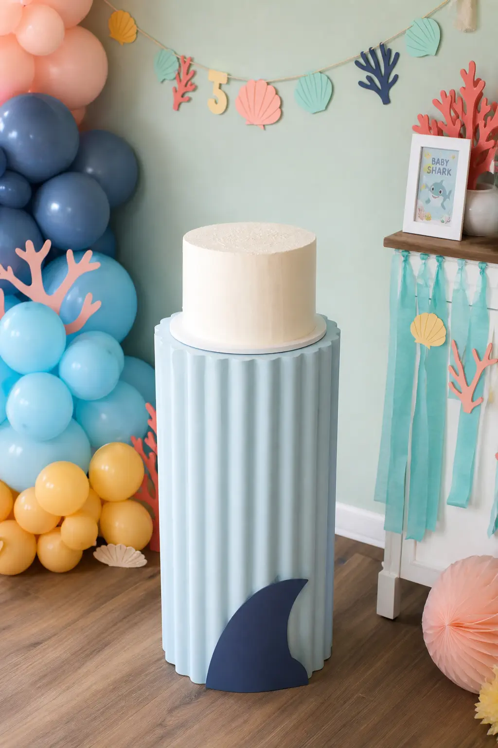 Fluted Shark Fin Cake Pedestal — A pale blue fluted pedestal with a single navy fin silhouette cut into its base does more theme work than any topper. Baby