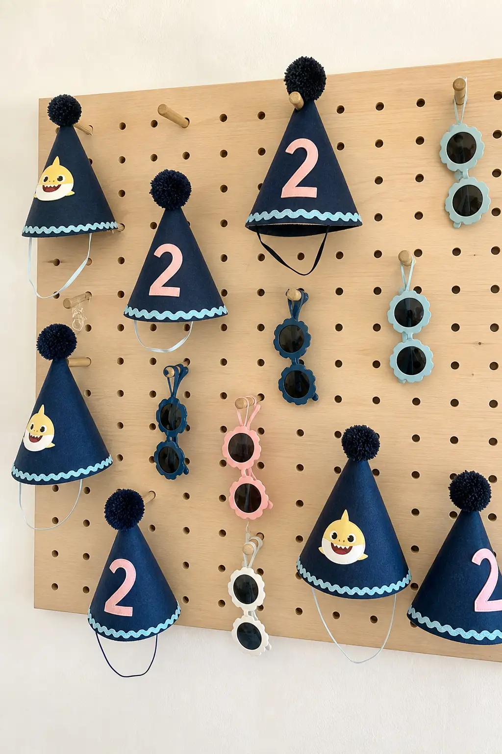 Party Hat and Sunglasses Pegboard — Navy pom-pom party hats and small round sunglasses hang from a plain wooden pegboard, letting guests pick their own on the