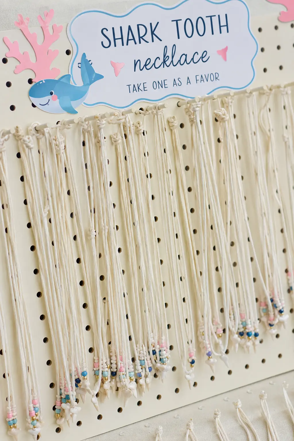 Shark Tooth Necklace Favor Board — Cream cord necklaces, each strung with one small tooth-shaped charm, hang in a row from a cream pegboard. Baby shark