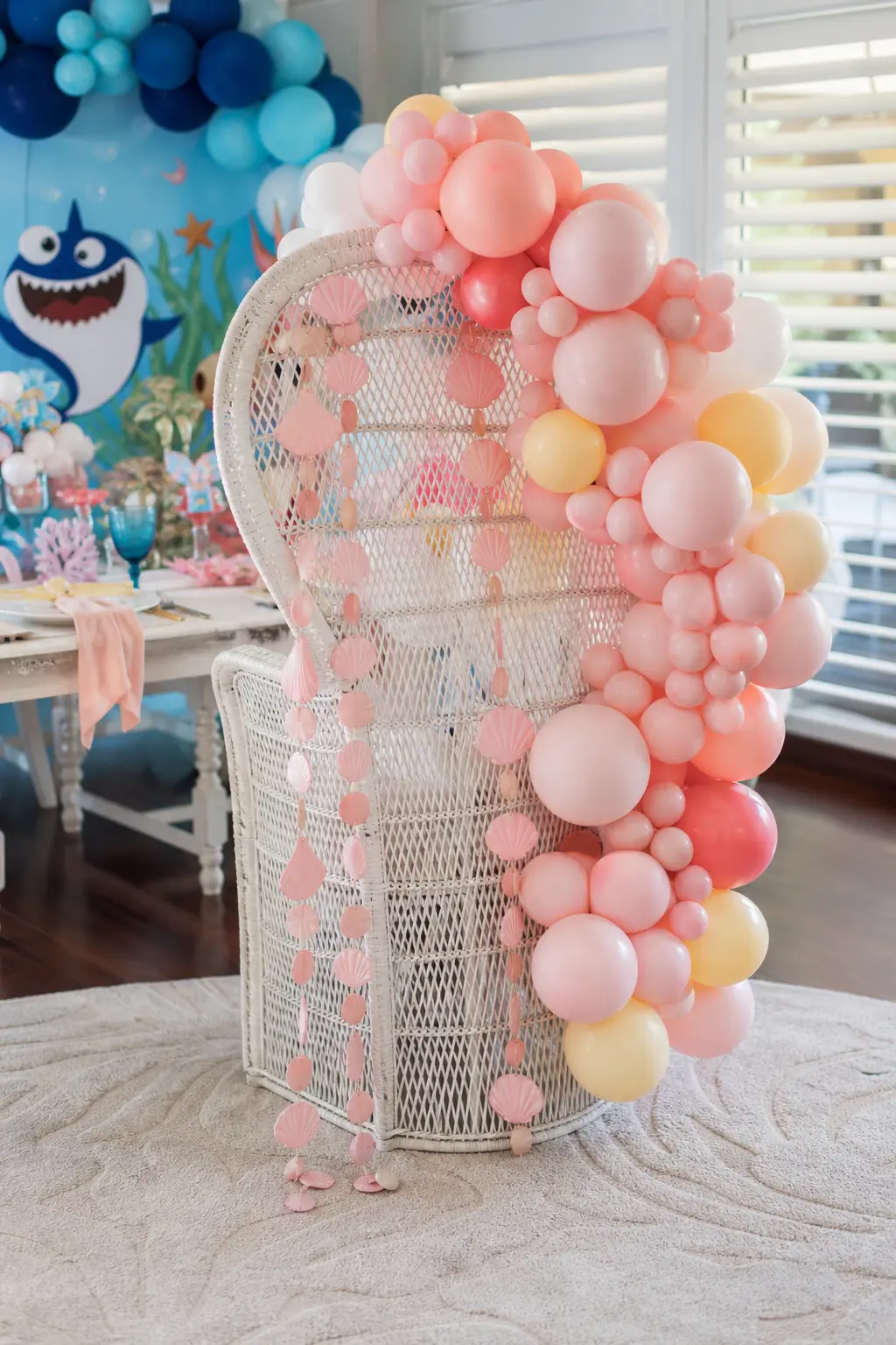 Shell and Ribbon Throne Chair — A white wicker chair, wrapped with blush balloon clusters and a trailing shell garland, gives the birthday girl a seat that