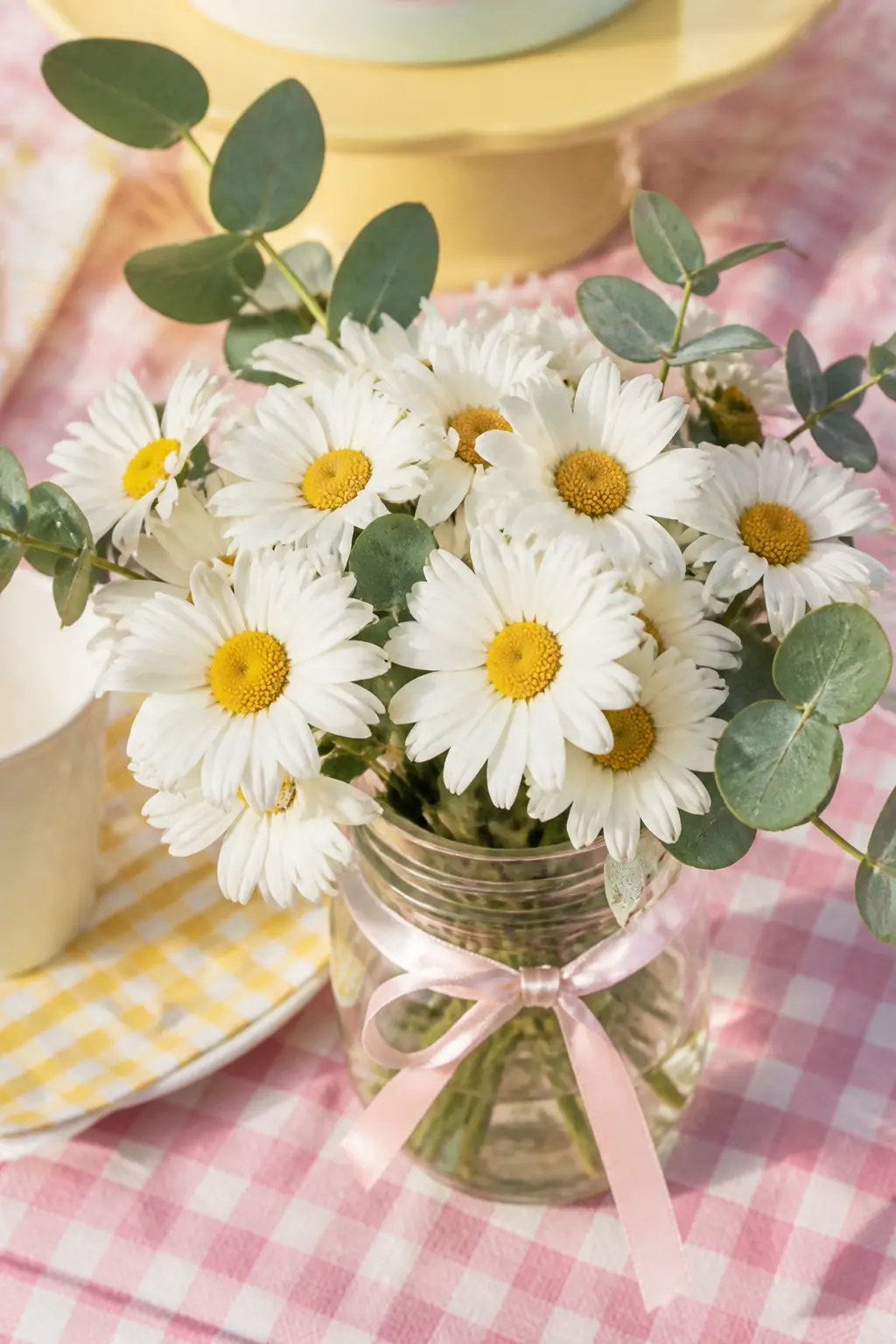 Daisy Centrepiece Posy — A jam jar filled with a tight posy of white daisies and a sprig of eucalyptus, tied with a narrow pink satin ribbon, repeating at