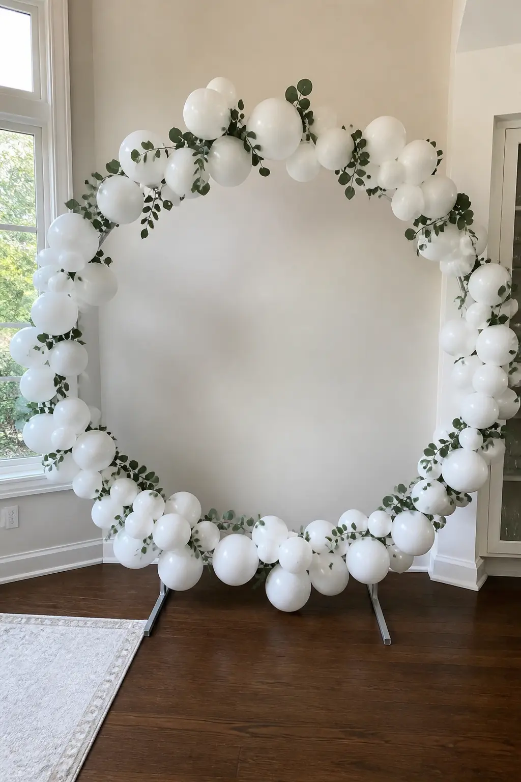 White Full Circle with Eucalyptus for Photographs — A full balloon circle in all white stands on two weighted bases in a light-filled corner. Bridal shower