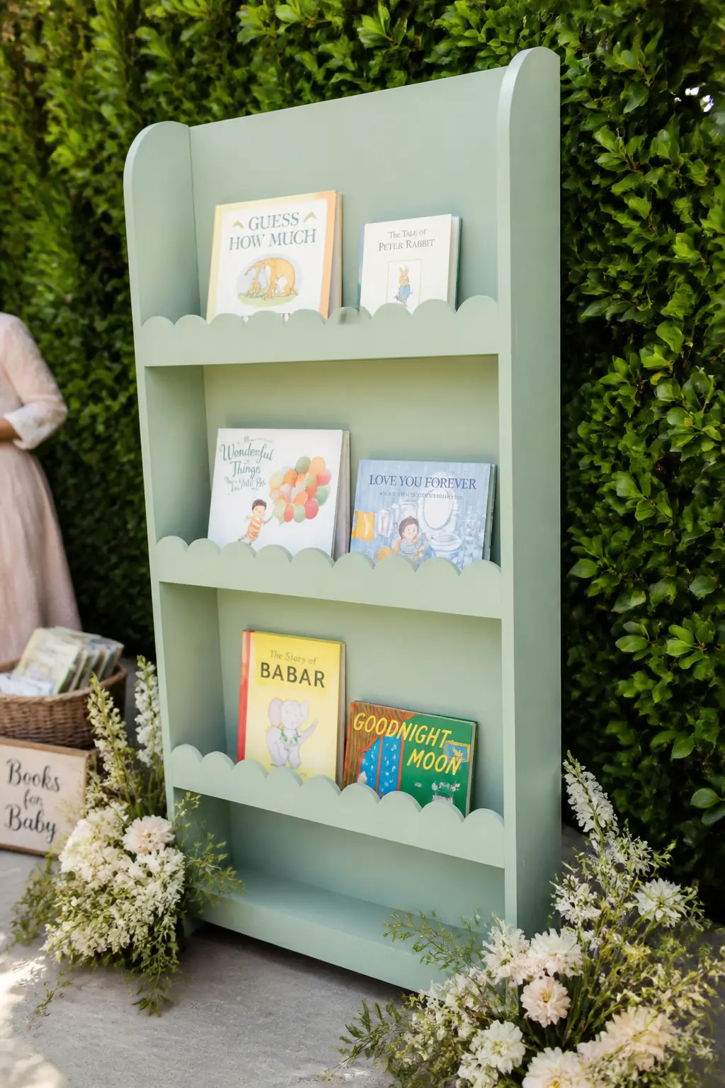 The Scallop-Edge Mint Bookshelf Backdrop — A freestanding bookshelf with a scalloped top edge, painted in a soft mint or duck-egg tone, placed against a hedge