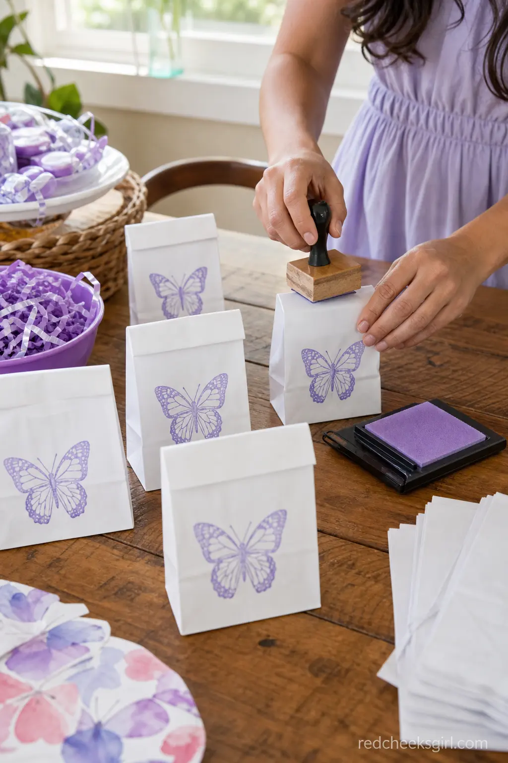 Butterfly-Stamped Favor Bag Station — A short stack of plain white paper bags, each stamped by hand with a lavender butterfly ink stamp, turns the cheapest