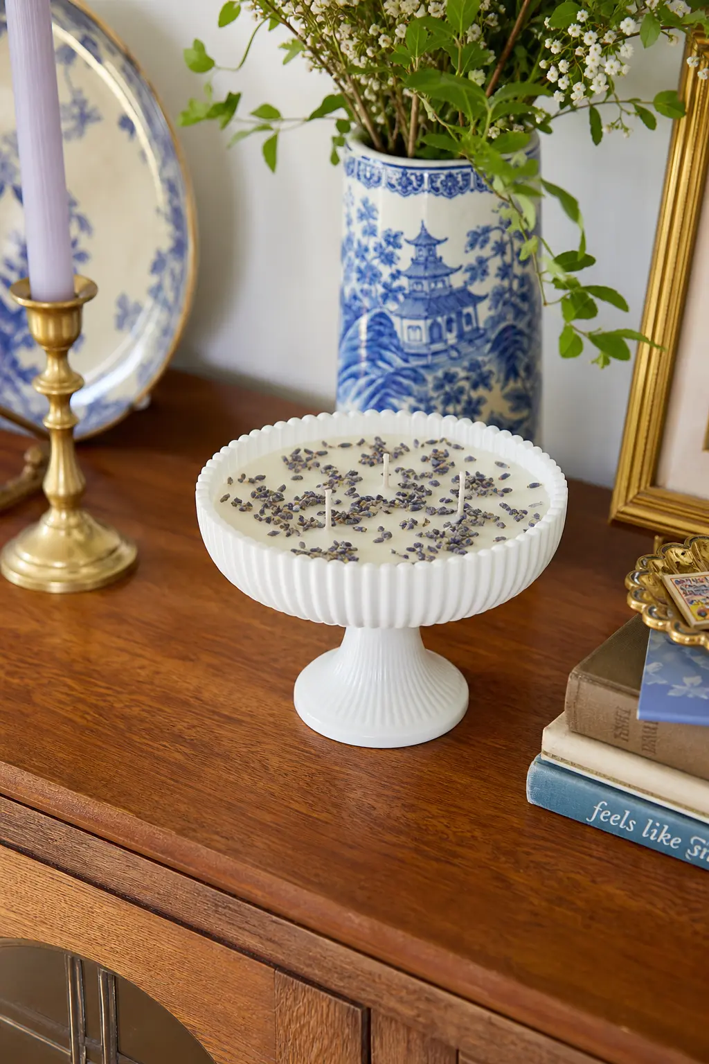 Milk Glass Compote Candle — A footed milk glass compote dish, opaque white and slightly ribbed, holds an ivory candle finished with a scattering of dried