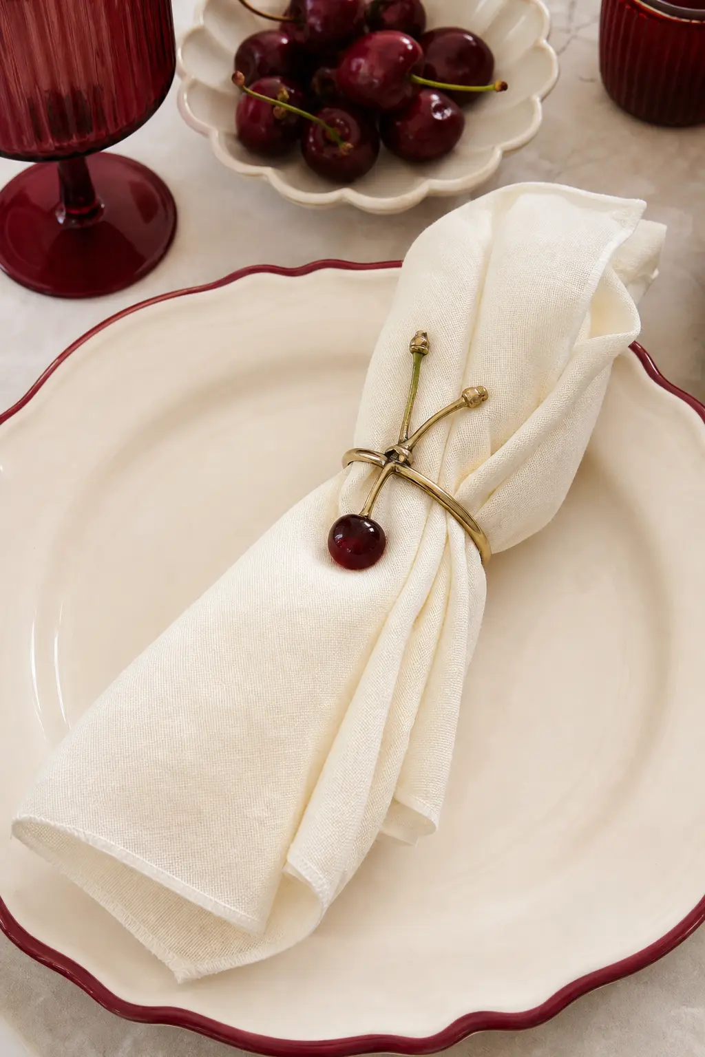 The Brass Cherry Stem Napkin Ring — A slim brass napkin ring holds a folded ivory napkin at each place, with one fresh cherry stem tucked through the ring