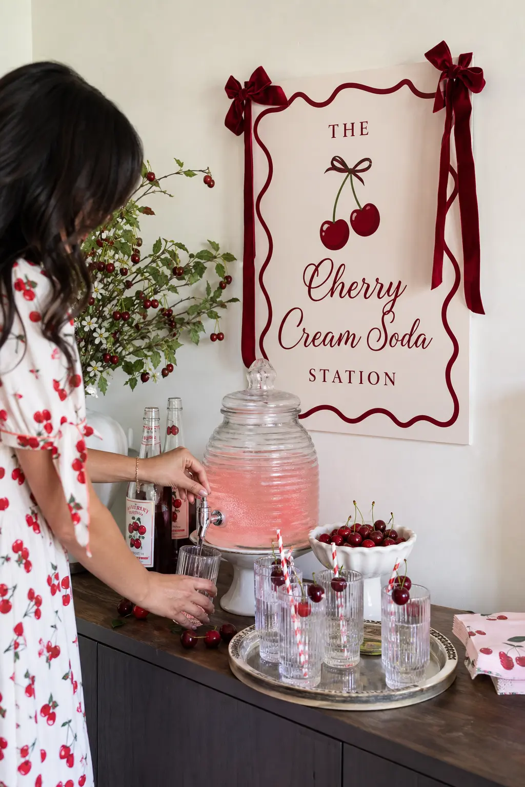 The Cherry Cream Soda Station — A glass beverage dispenser holds a pale pink cherry cream soda, with a small tray of glasses and fresh cherries alongside for