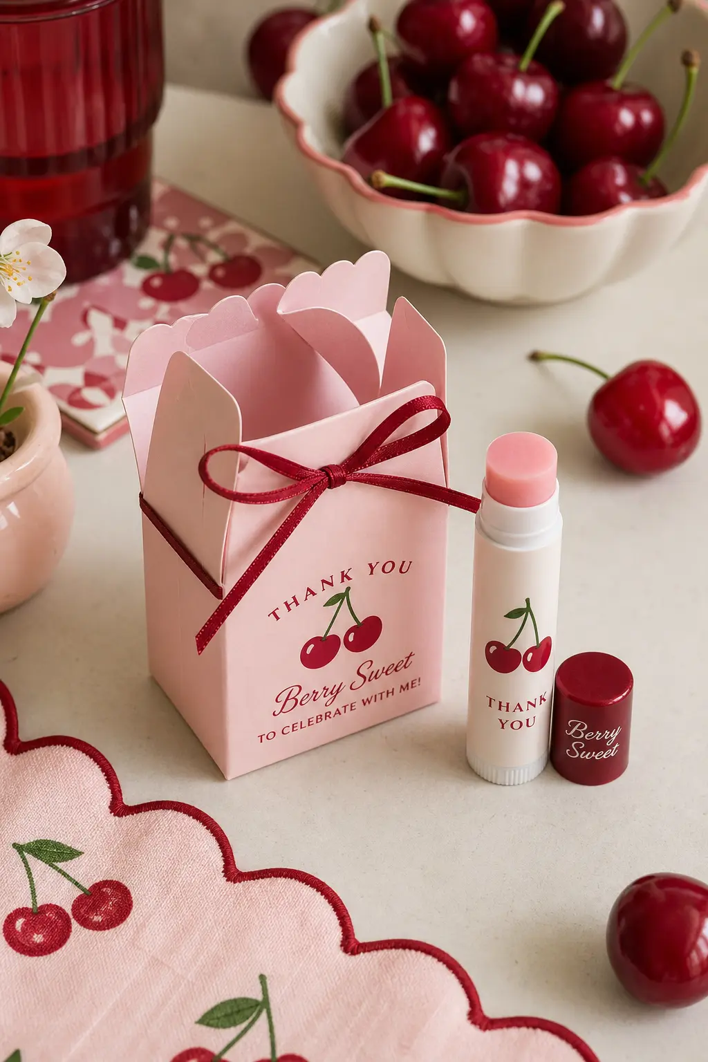 The Cherry Lip Balm Favor Box — A small blush pink box holds a cherry flavored lip balm, tied shut with a thin cherry red ribbon and nothing printed on the lid