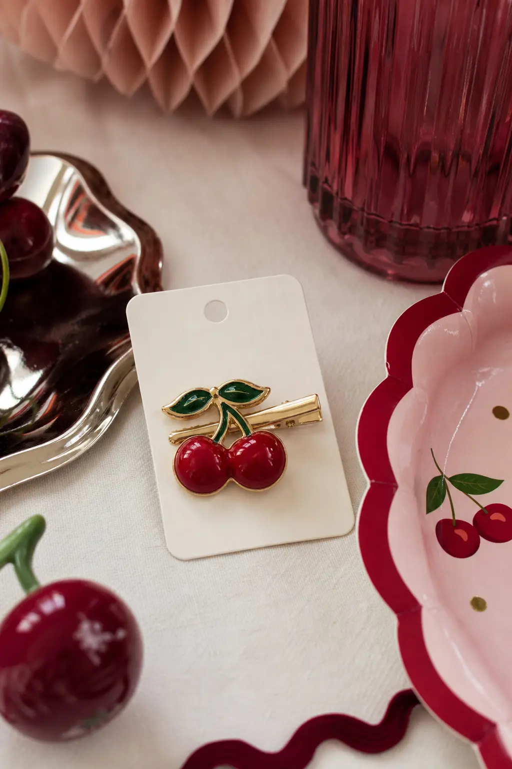 The Enamel Cherry Hair Clip Card — A small enamel cherry hair clip sits pinned to a plain cream card, no printed text, just the fruit shape itself in glossy