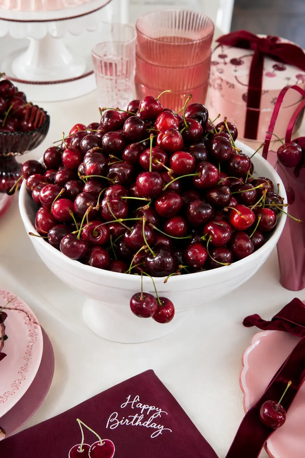 The Overflowing Cherry Bowl — A wide, glossy white ceramic bowl piled generously with fresh cherries sits at the center of the table, doing more work than any