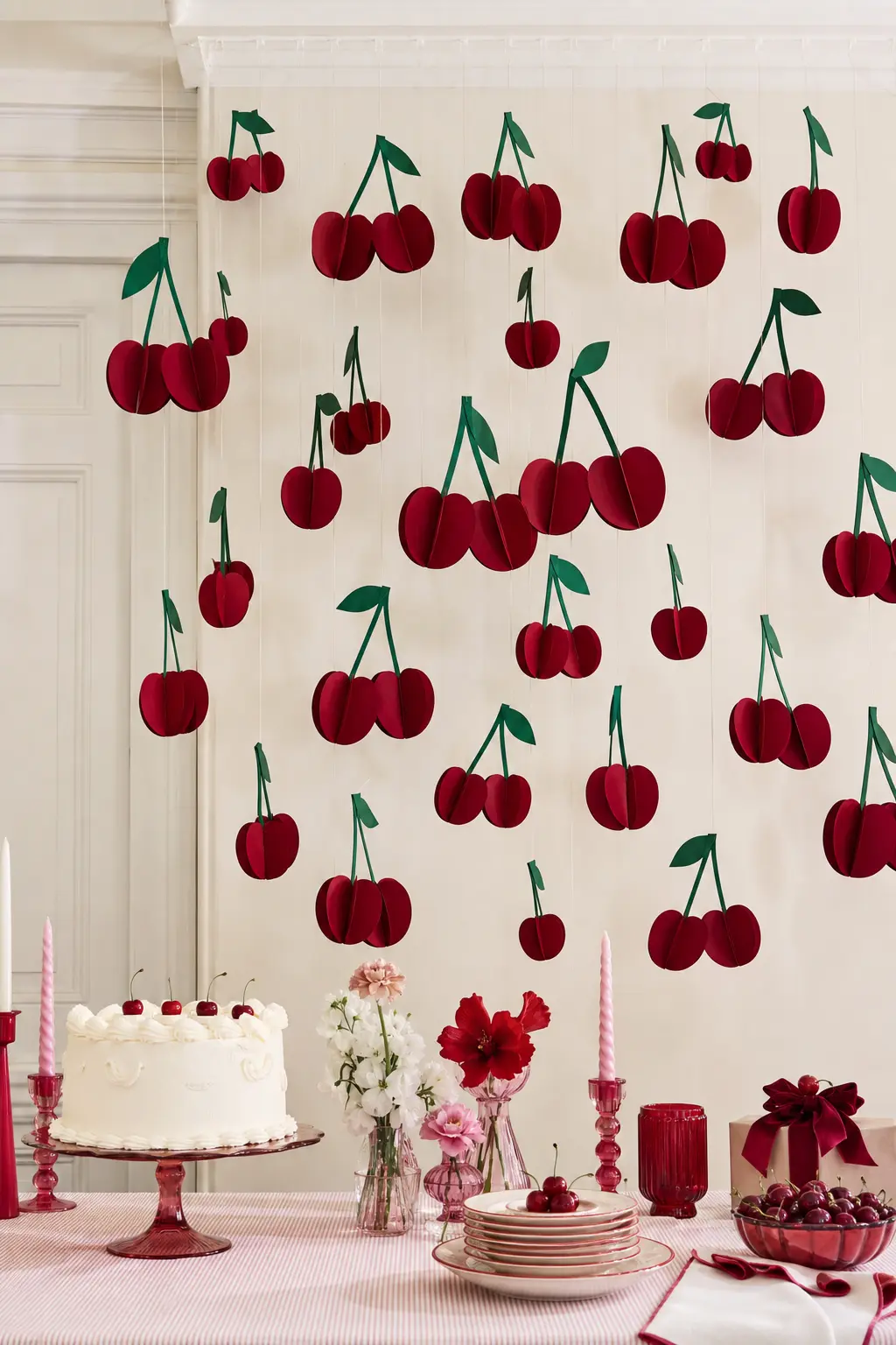The Paper Cherry Cloud Wall — Dozens of oversized paper cherries in deep red and forest green hang at different lengths from the ceiling, forming a loose cloud