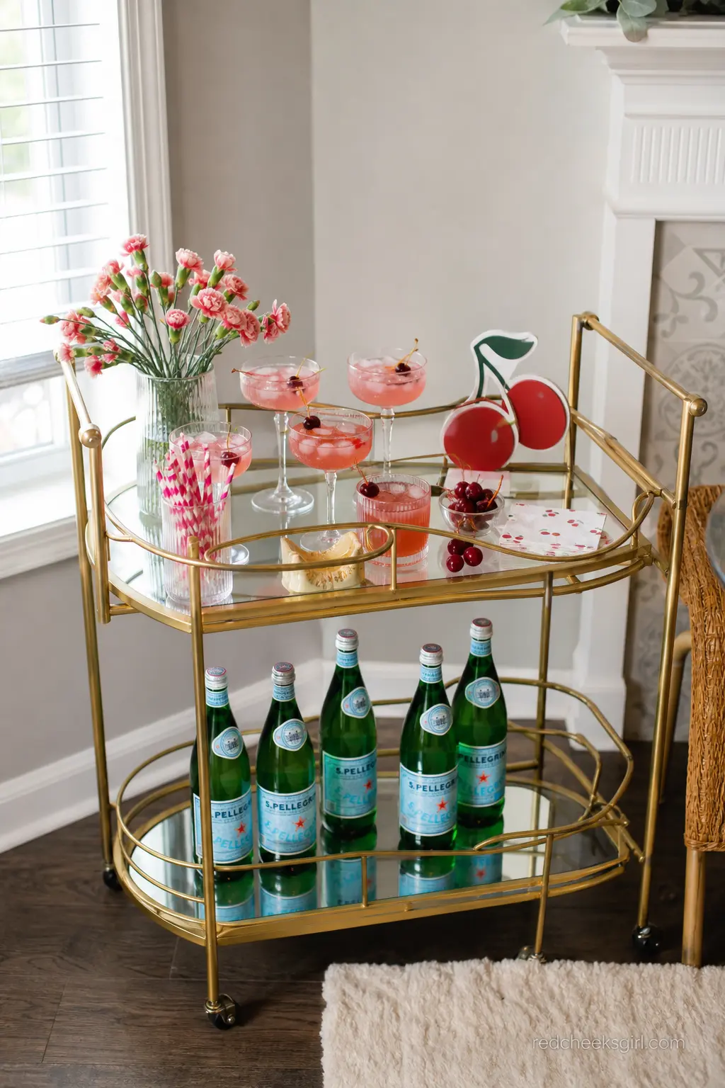 Brass Cart Cherry Mocktail Bar — A small brass or gold bar cart holding pink mocktails garnished with fresh cherries turns a corner of the living room into its