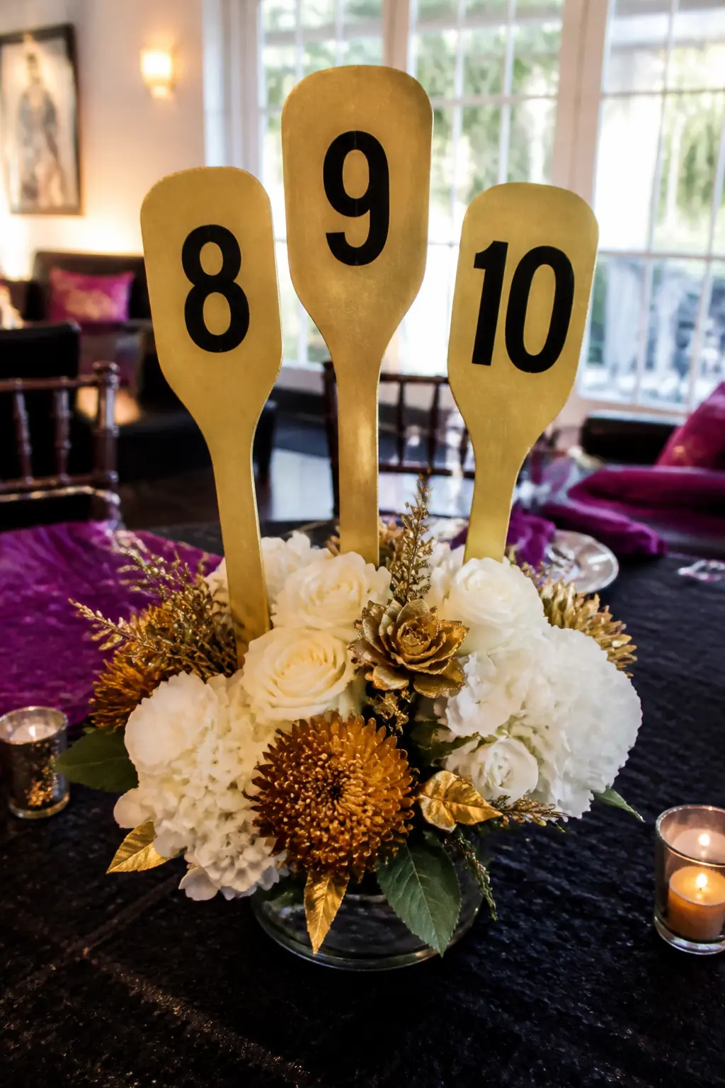 Paddle Centrepiece With Numbered Cards — Three gold-painted wooden paddles, each showing a number from 8 to 10, stand in a low vase or a block of floral foam