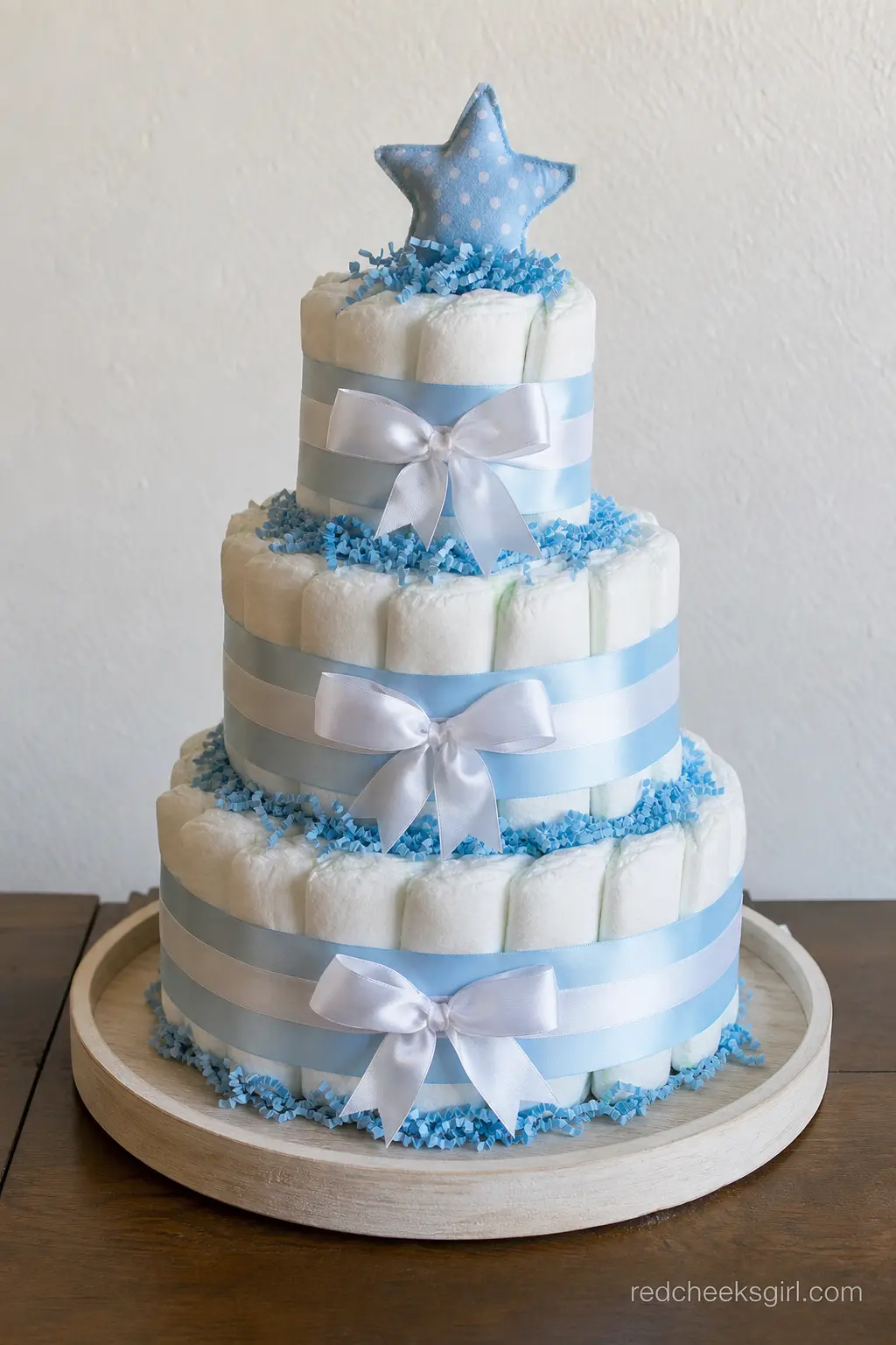Classic Three-Tier Blue Ribbon Diaper Cake — Three tiers of rolled diapers wrapped in baby blue satin, finished with a wide white bow and a puff of tulle at
