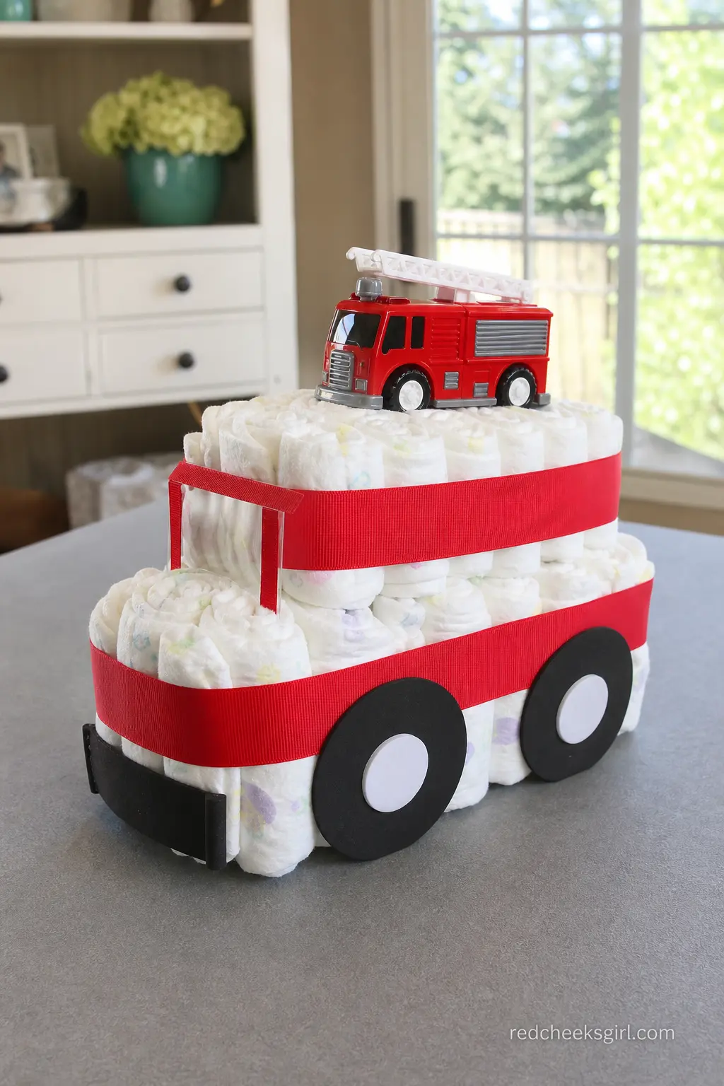 Fire Truck Diaper Cake — Bright red ribbon wraps the tiers, black felt circles stand in for wheels along the base, and a die cast fire truck sits on top like