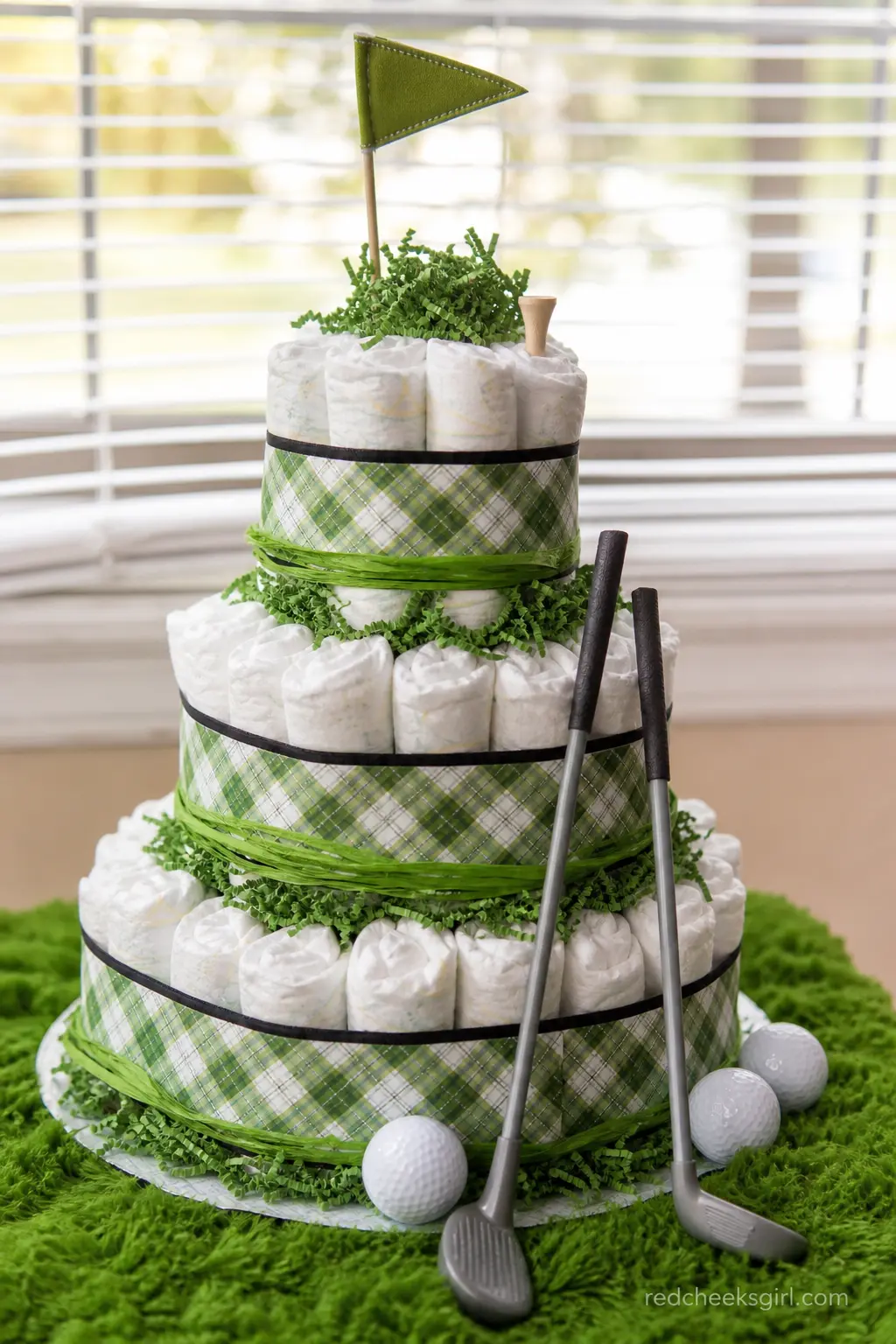 Golf Diaper Cake — Green and white plaid ribbon wraps the base, a scattering of mini golf balls sits between tiers, and a set of tiny toy clubs leans against